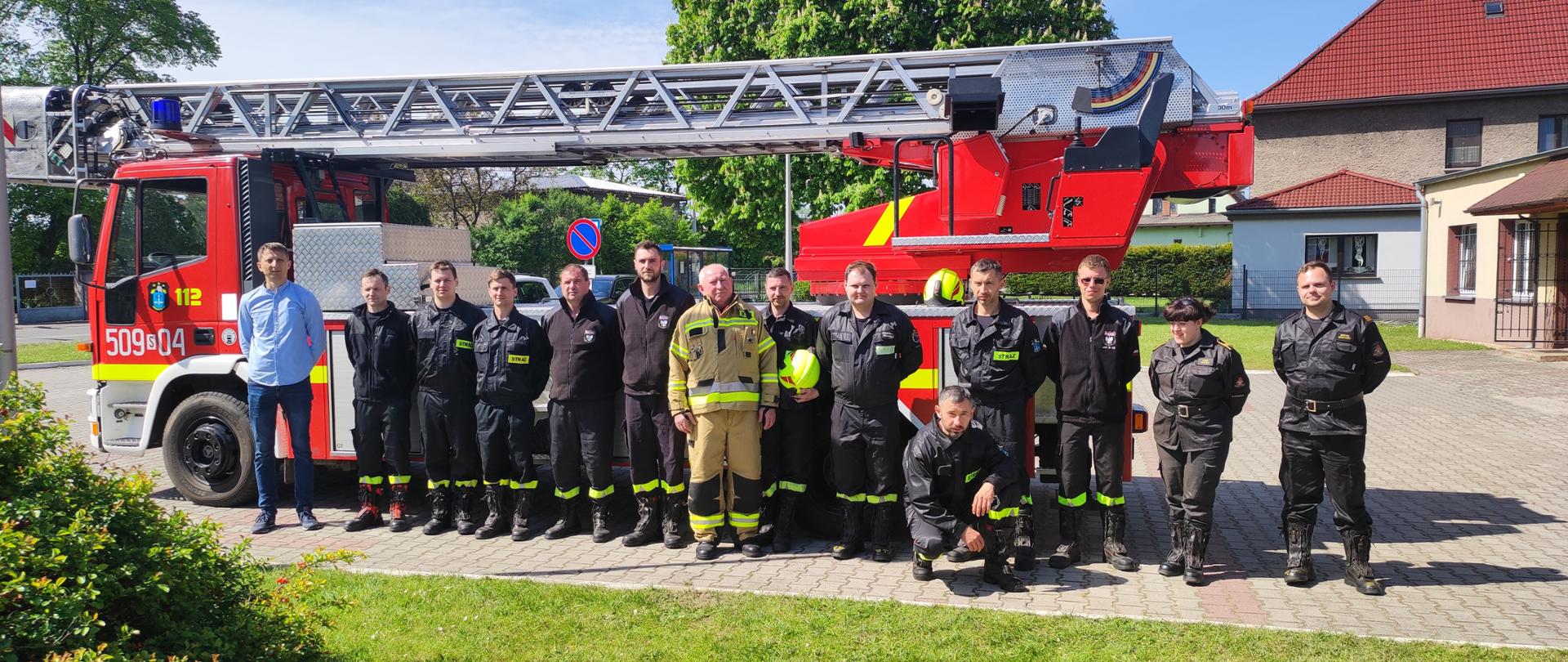 na zdjęciu stoi na placu OSP grupa strażaków na tle samochodu drabiny mechanicznej