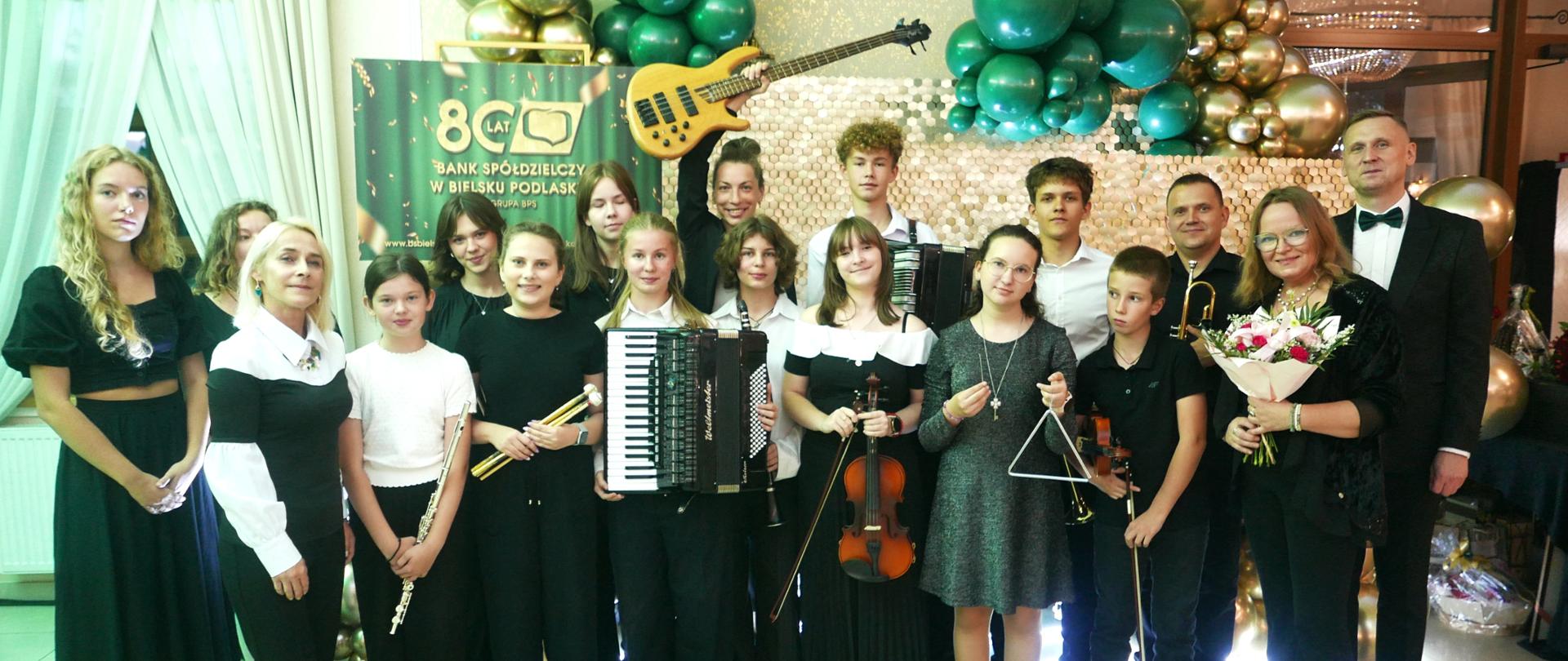 Młodzież i dorośli z zespołu pozują do zdjęcia z Prezesem Banku. Niektórzy z nich trzymają instrumenty muzyczne. W tle napis 80 Lat Banku Spółdzielczego w Bielsku Podlaskim, widzimy też złote i zielone baloniki 