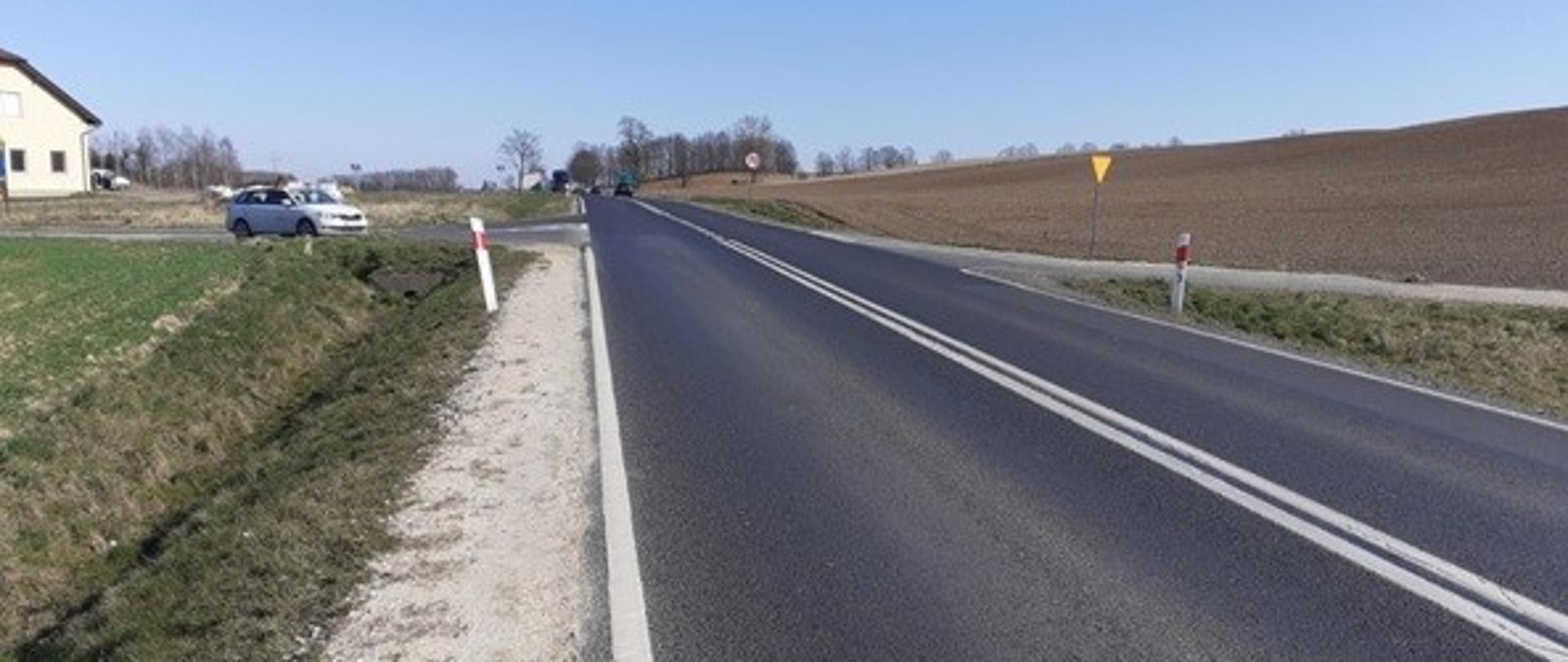 Skrzyżowanie dwóch dróg. Po lewej stronie zabudowa mieszkaniowa, po prawej pola uprawne.