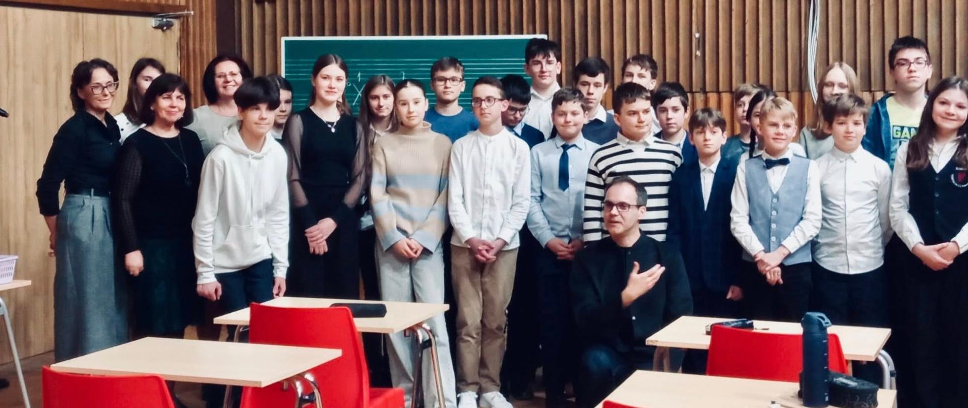 Grupa uczniów i nauczycieli - uczestników I Makroregionalnego Konkursu Kształcenia Słuchu pozuje wspólnie do zdjęcia w sali lekcyjnej. W tle widać tablicę i drewniane ściany, a na pierwszym planie ustawione są stoły i czerwone krzesła. Wszyscy stoją blisko siebie, uśmiechając się do obiektywu, co tworzy ciepłą i przyjazną atmosferę.