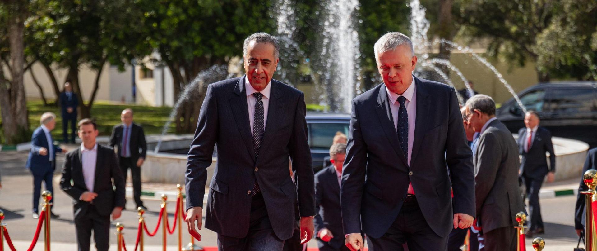 Minister Tomasz Siemoniak meets with Abdellatif Hammouchi, Director General of Internal Security of Morocco. In the foreground, on the right, is Minister Tomasz Siemoniak, and on the left, Abdellatif Hammouchi, Director General of Internal Security of Morocco. The ministers walk up the stairs on a red carpet. To the right and left, golden posts with a red rope. The posts separate the carpet from the rest of the area. Other people are visible to the right, left, and behind the ministers. In the background, a fountain, cars, trees, and buildings.