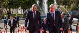 Minister Tomasz Siemoniak meets with Abdellatif Hammouchi, Director General of Internal Security of Morocco. In the foreground, on the right, is Minister Tomasz Siemoniak, and on the left, Abdellatif Hammouchi, Director General of Internal Security of Morocco. The ministers walk up the stairs on a red carpet. To the right and left, golden posts with a red rope. The posts separate the carpet from the rest of the area. Other people are visible to the right, left, and behind the ministers. In the background, a fountain, cars, trees, and buildings.