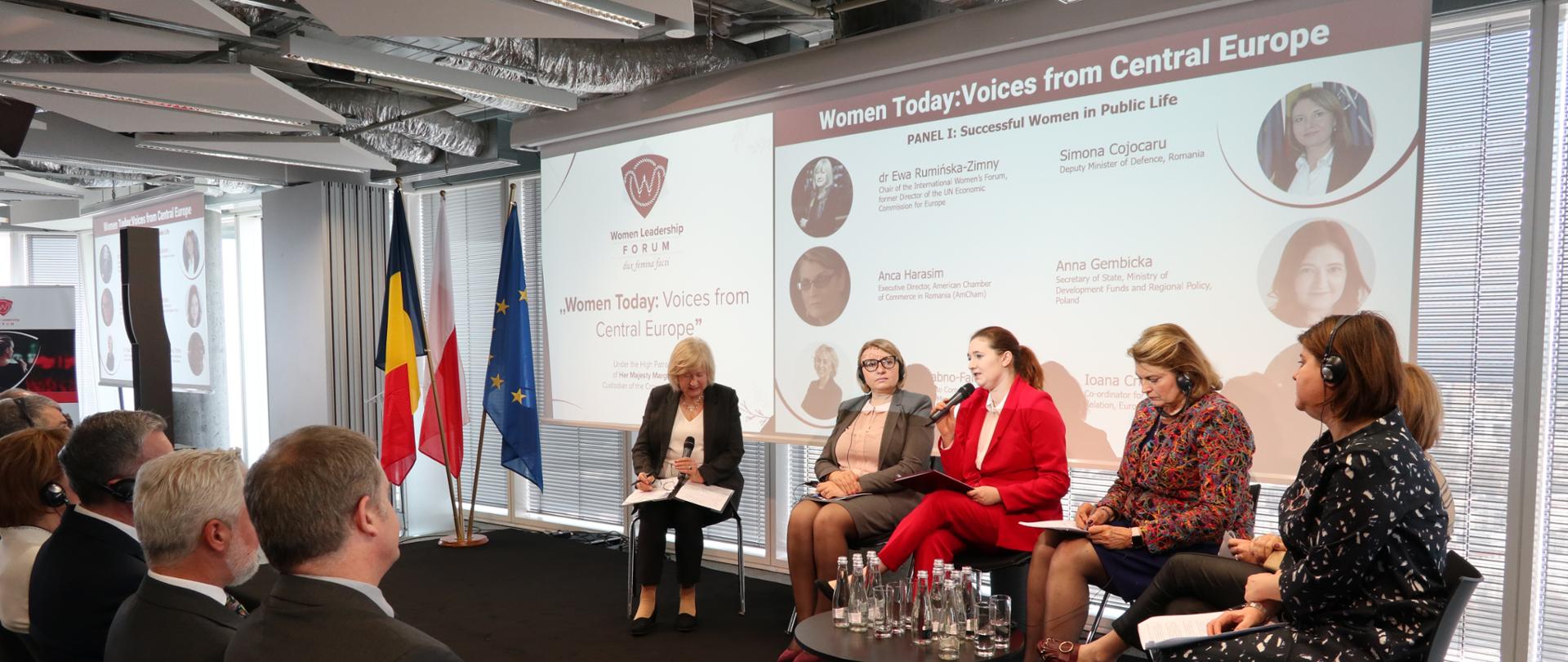 Wokół stolika siedzą paneliści, publiczność patrzy w stronę sceny, za panalistkami wyświetlana jest prezentacja