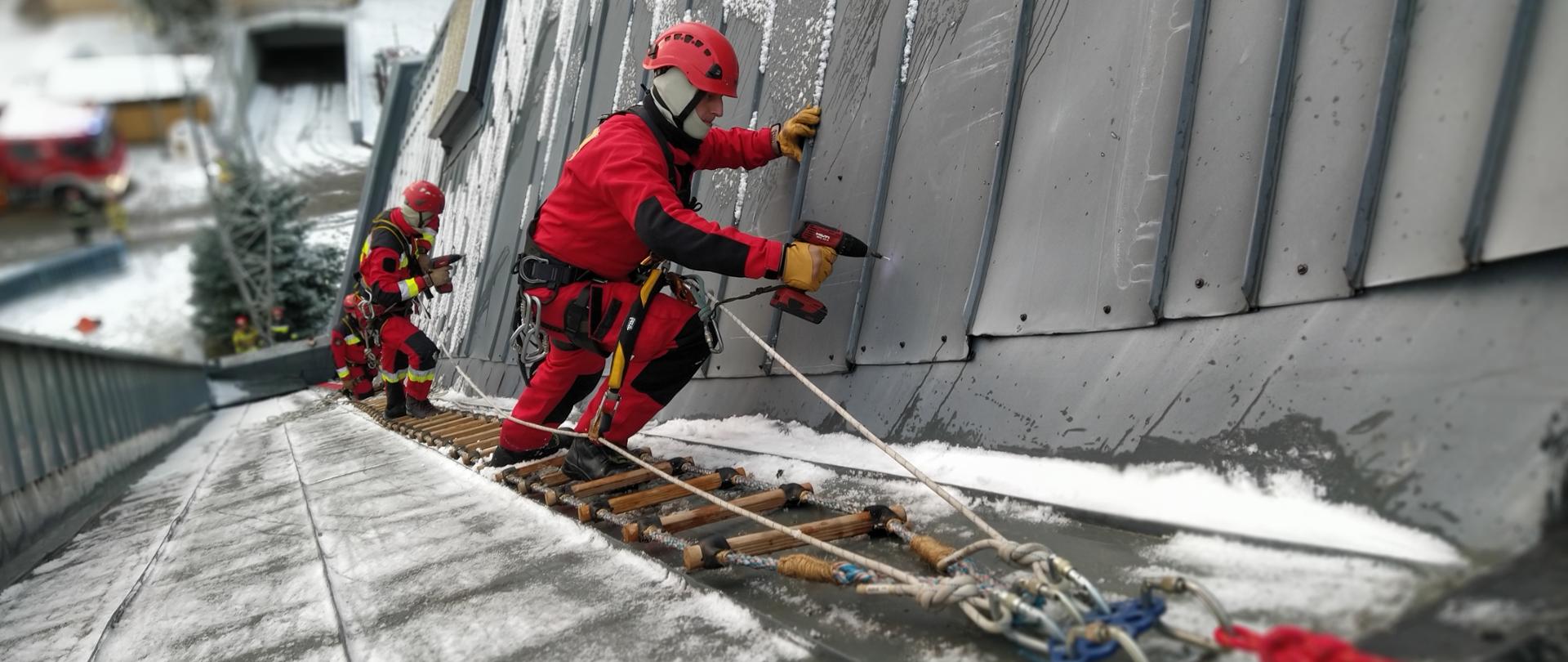 Zdjęcia przedstawiają ratowników Grupy Wysokościowej znajdujących się na dachu hali widowiskowej Podpromie. Ratownicy są zabezpieczeni specjalistycznym sprzętem przed upadkiem z wysokości. Za pomocą wkrętarek i wkrętów zabezpieczają oderwaną blachę na dachu obiektu. 