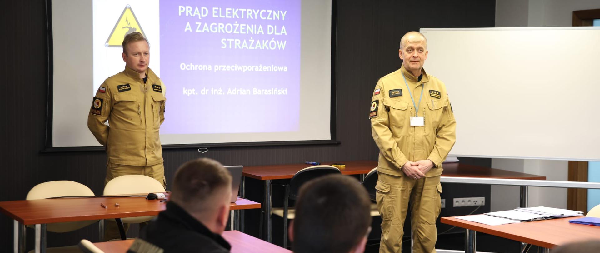 Dwóch oficerów CS PSP stoi na środku sali w pewnej odległości od siebie, za nimi rozwinięty ekran multimedialny z wyświetlonym slajdem prezentacji