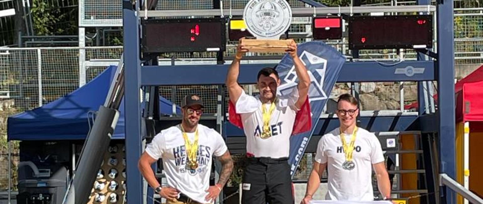 Trzech funkcjonariuszy stojących na podium zawodów, w tle wieża zawodów.