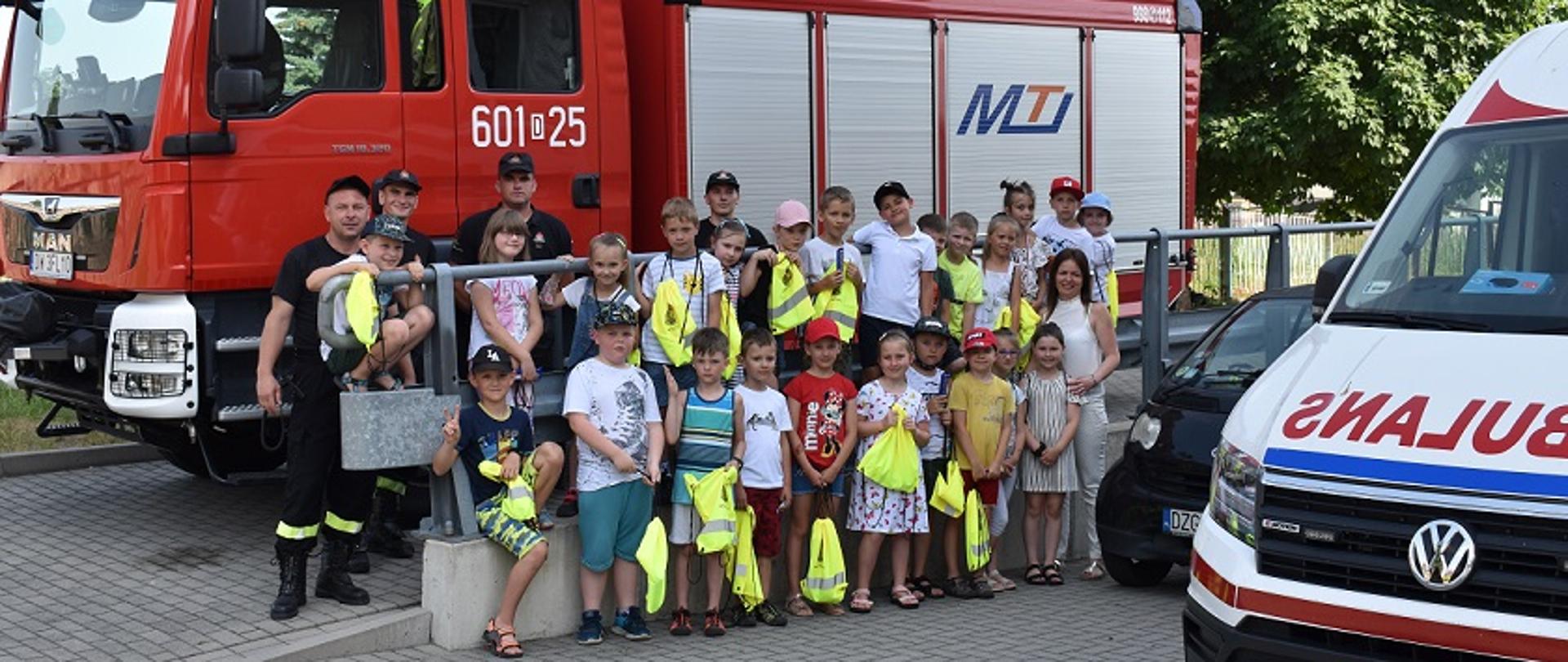 Grupka dzieci stoi na tle samochodu ratowniczo-gaśniczego o numerze operacyjnym 601D25. Między dziećmi stoją strażacy. Z prawej strony widać część przodu karetki pogotowia.