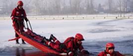 Strażacy ubrani w kombinezony do pracy w wodzie podczas podejmowania osoby poszkodowanej z zarwanego lodu na deskę lodową