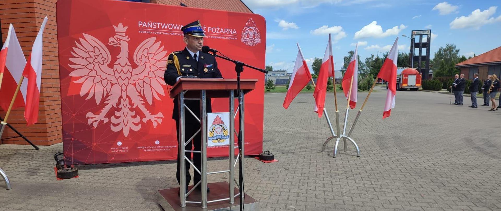 przemówienie wielkopolskiego komendanta wojewódzkiego PSP w tle flagi biało czerwone oraz czerwona ścianka z orłem 