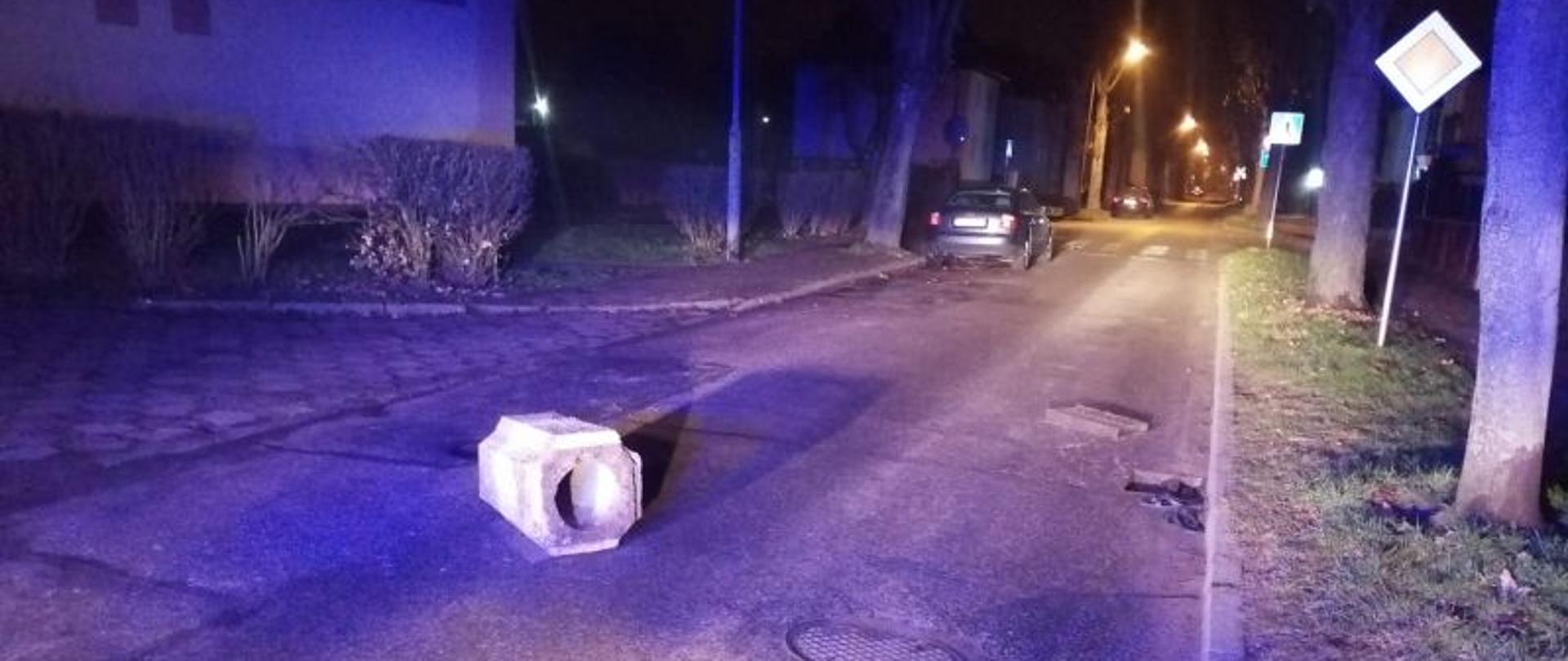 Zdjęcie wykonane w nocy. Widok na długą ulicę. Ulica oświetlona niebieskimi reflektorami. Na środku drogi betonowy śmietnik.