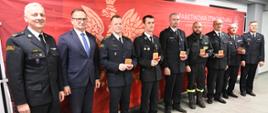 Strażacy wraz z prezydentem miasta stoją w szeregu przed czerwonym tłem z logo i napisem „Państwowa Straż Pożarna”. Większość z nich ma na sobie galowe mundury strażackie, a jeden z nich jest w roboczym ubraniu strażaka. Każdy trzyma w dłoni czerwone etui z odznaczeniem. Wszyscy patrzą w stronę aparatu i pozują do pamiątkowego zdjęcia.