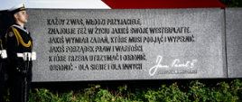 Żołnierz stoi obok pomnika na którym widnieje napis "Każdy z Was, młodzi przyjaciele, znajduje też w życiu jakieś swoje Westerplatte. Jakiś wymiar zadań, które musi podjąć i wypełnić. Jakiś porządek praw i wartości, które trzeba utrzymać i obronić, obronić - dla siebie i dla innych" Jan Paweł II Westerplatte, 12 czerwca 1987 pod pomnikiem rosną krzewy oraz trawa. 