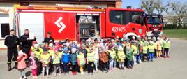 Zdjęcie grupowe dzieci ze strażakami. W tle samochody strażackie.
