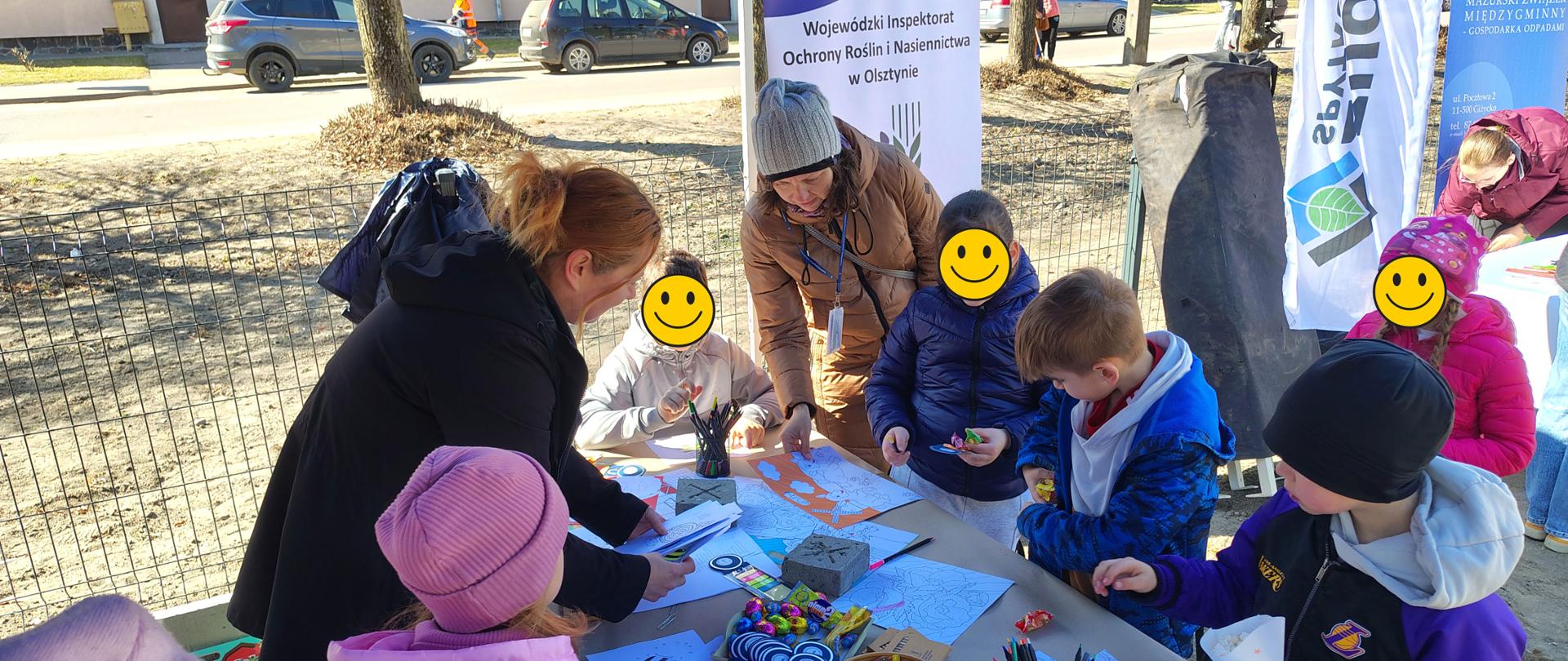 na stole rozłożone kolorowanki, kredki, cukierki, nad nimi pochylona grupka dzieci z dorosłymi, w tle baner WIORiN