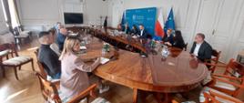 Grupa osób przy stole konferencyjnym w tle niebieska plansza z napisem Wojewoda Małopolski, w tle również flagi: polska i Unii Europejskiej