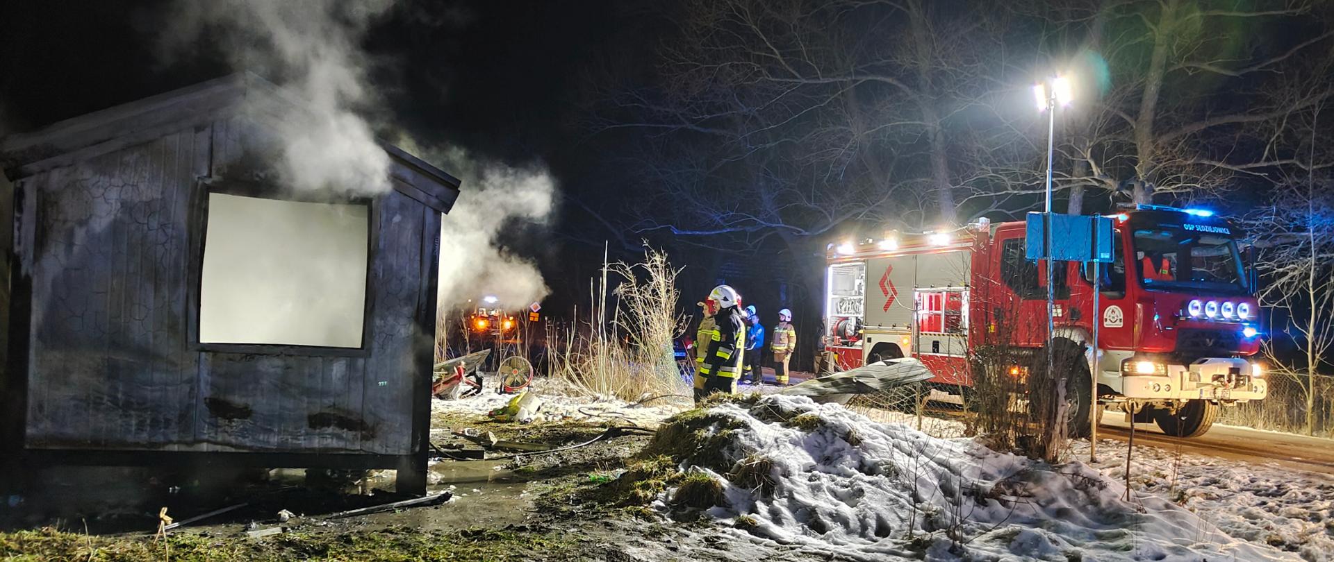 Na zdjęciu po lewej stronie widać spalony kontener letniskowy, po prawej zaś samochód pożarniczy. W centralnej części fotografii widać ratownika, a w głębi na nim innych strażaków.