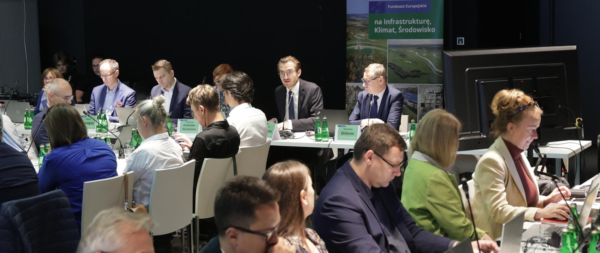 Grupa osób siedzi przy stołach w sali konferencyjnej