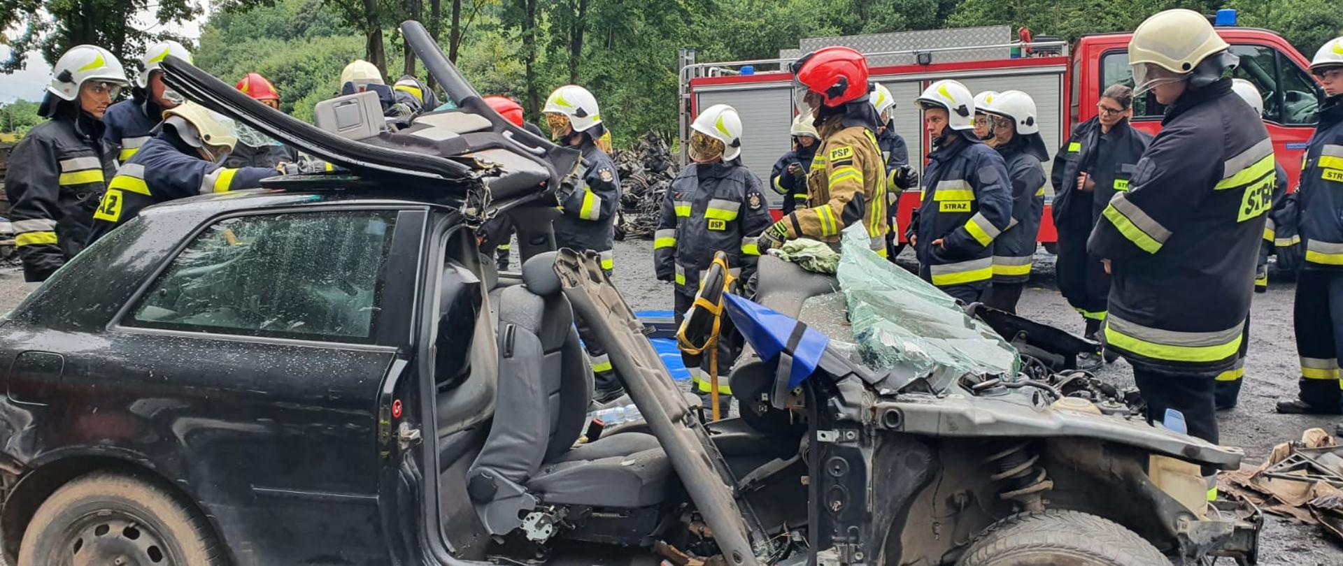 Zdjęcie przedstawia grupę strażaków podczas szkolenia podstawowego. Strażacy ćwiczą cięcie, rozpieranie elementów pojazdów. Zajęcia z tematyki: metody uwalniania osób poszkodowanych z samochodów osobowych, ciężarowych oraz autobusów – zajęcia przeprowadzane w punkcie kasacji pojazdów Eco-Auto-System w Rąbinie.