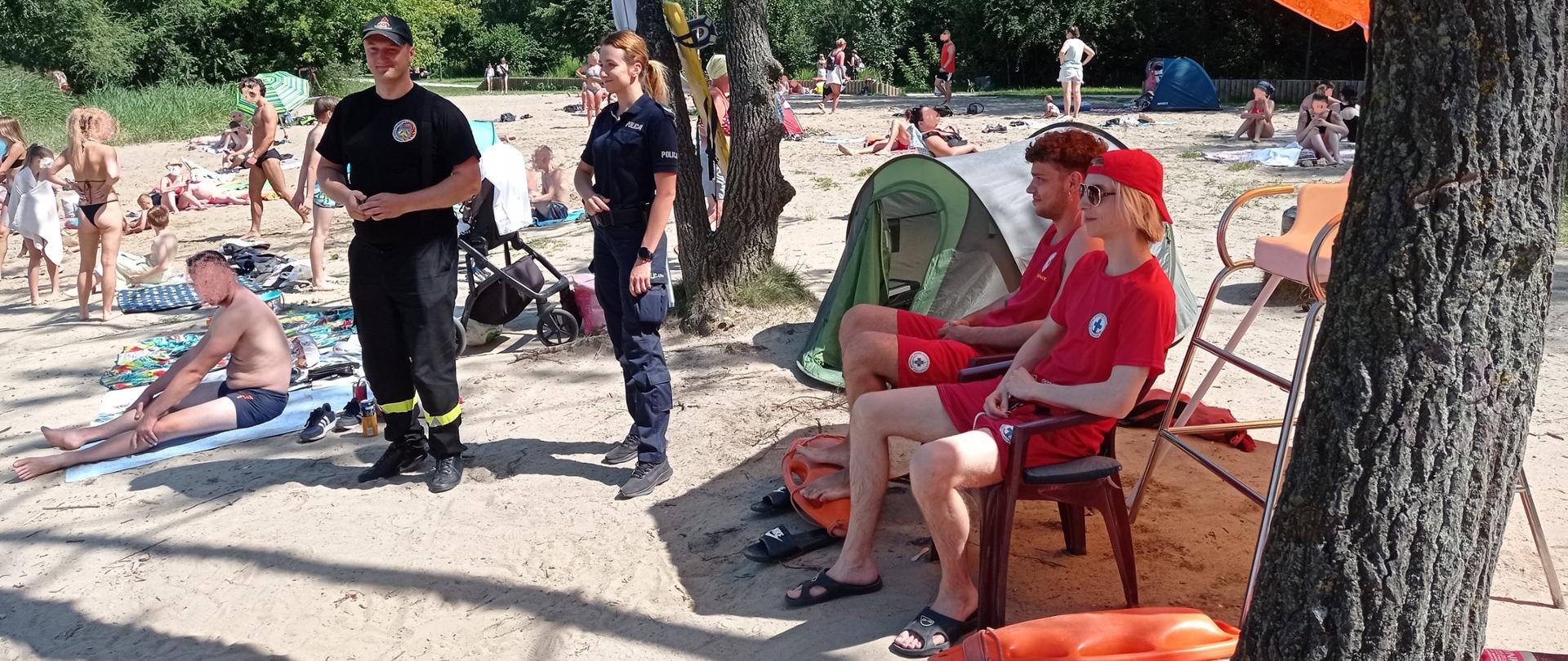 Zdjęcie przedstawia piaszczystą plażę na której rośnie kilka drzew. Na pierwszym planie widać ratowników wodnych ubranych w czerwone koszulki i spodenki. Obok stoi policjantka w granatowym uniformie i strażak w czarnym uniformie. Na drugim planie widać zażywających słonecznych kąpieli ludzi.