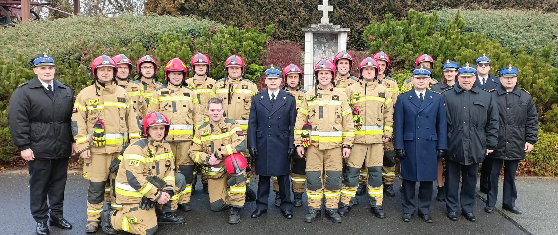 Na tle kapliczki i roślinności na skarpie ustawieni strażacy do wspólnego zdjęcia. W środku pomiędzy dwoma oficerami w umundurowaniu wyjściowym stoją przyszli emeryci. Strażacy zmiany służbowej w ubraniach specjalnych. Strażacy systemu codziennego w umundurowaniu wyjściowym – kurtkach lub płaszczach oraz rogatywkach. 