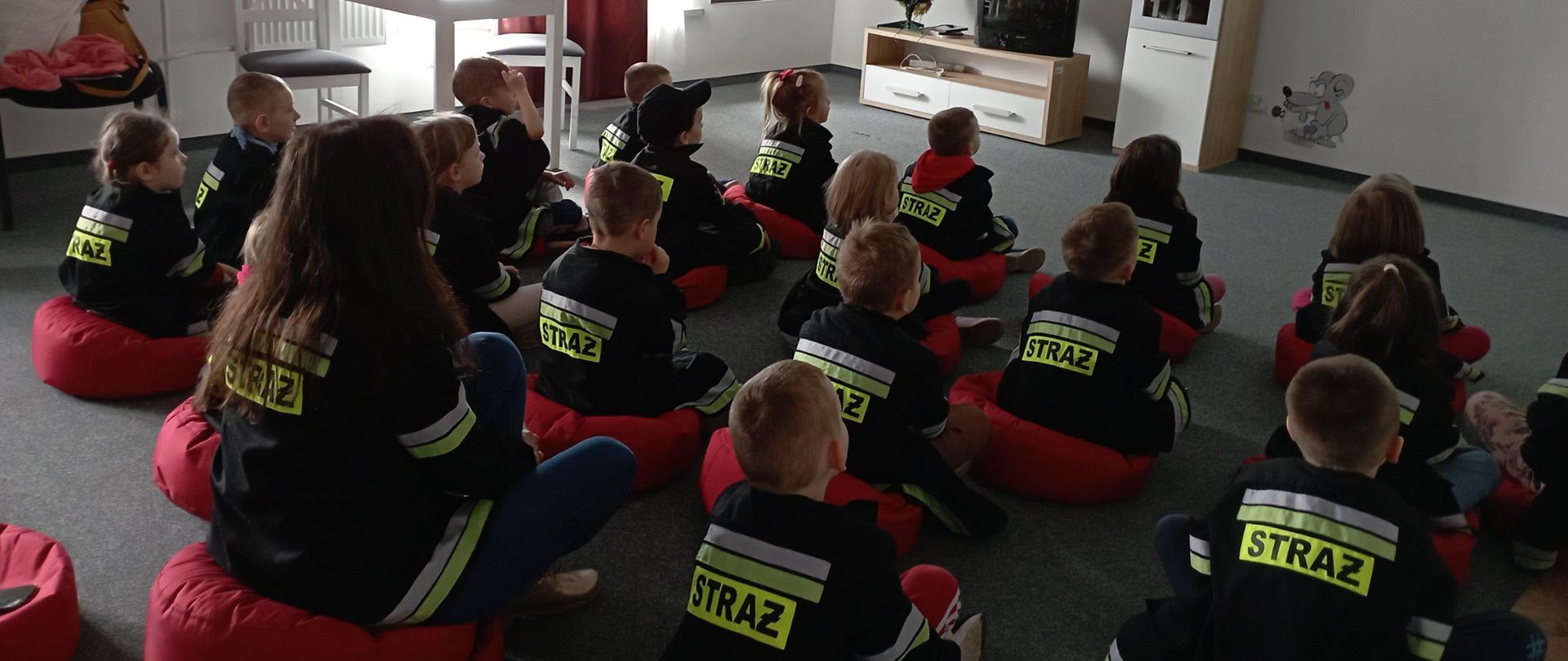 Na zdjęciu przedstawiono zajęcia w sali Ognik. Dzieci w przebraniu strażaka siedząc wraz z opiekunem na podłodze oglądają film edukacyjny.