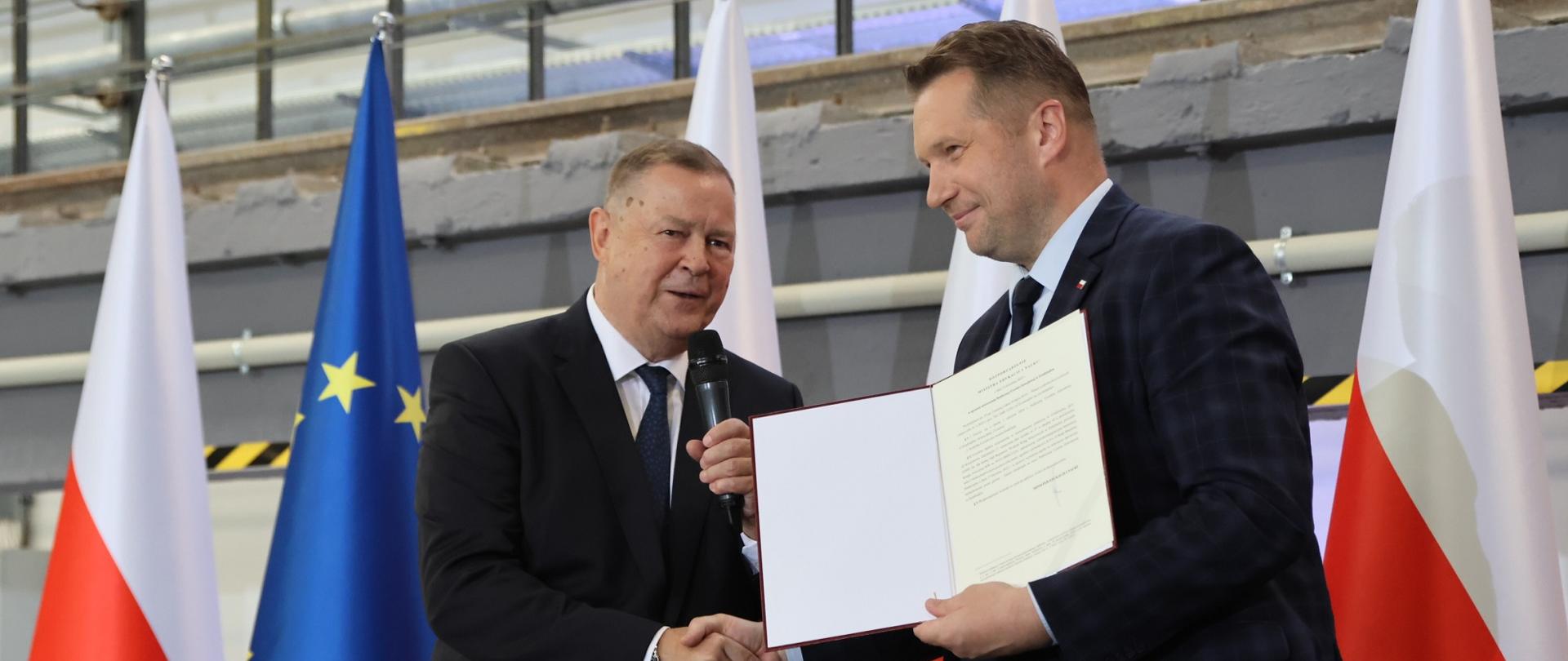 Minister Czarnek trzyma otwartą teczkę, ściska rękę stojącemu obok mężczyźnie w garniturze, za nimi kilka polskich flag.
