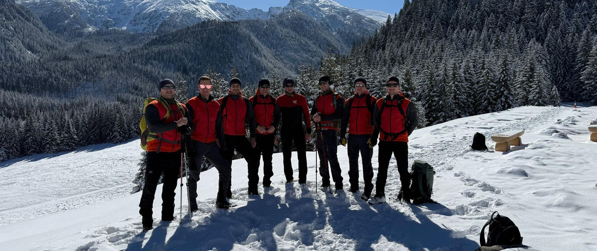 Grupa siedmiu osób w czerwonych kurtkach stoi na ośnieżonej górze z kijkami narciarskimi. Ośnieżone szczyty i lasy są widoczne pod czystym, błękitnym niebem.
