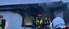 Strażacy w aparatach powietrznych przed budynkiem, wydobywa się dym z pomieszczenia