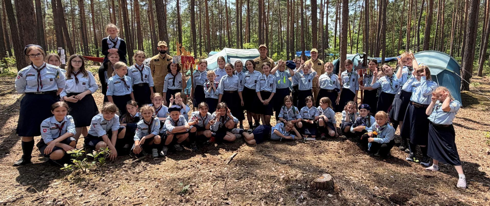 Zdjęcie przedstawia dużą grupę harcerek (zuchów lub harcerek starszych) oraz trzech strażaków w beżowych mundurach, pozujących wspólnie w lesie iglastym. Dziewczęta są ubrane w oficjalne mundury: jasnoniebieskie koszule z ciemnymi chustami oraz ciemne spódnice. Część grupy stoi, część klęczy lub siedzi bezpośrednio na leśnej ściółce. Uczestniczki uśmiechają się, niektóre wykonują wesołe gesty do aparatu. W tle widać rozbite turystyczne namioty oraz gęsty las z wysokimi, prostymi pniami słońca. Na pierwszym planie widoczny jest fragment pnia drzewa. Słoneczny dzień, światło przenika przez korony drzew.