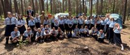Zdjęcie przedstawia dużą grupę harcerek (zuchów lub harcerek starszych) oraz trzech strażaków w beżowych mundurach, pozujących wspólnie w lesie iglastym. Dziewczęta są ubrane w oficjalne mundury: jasnoniebieskie koszule z ciemnymi chustami oraz ciemne spódnice. Część grupy stoi, część klęczy lub siedzi bezpośrednio na leśnej ściółce. Uczestniczki uśmiechają się, niektóre wykonują wesołe gesty do aparatu. W tle widać rozbite turystyczne namioty oraz gęsty las z wysokimi, prostymi pniami słońca. Na pierwszym planie widoczny jest fragment pnia drzewa. Słoneczny dzień, światło przenika przez korony drzew.