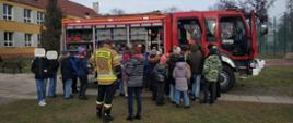 Na terenie Szkoły Podstawowej nr 2 w Łowiczu grupa dzieci ogląda wyposażenie samochodu ratowniczo gaśniczego barwy czerwonej w samochodzie otwarte skrytki ze sprzętem otwarte drzwi w kabinie pojazdu niebo bezchmurne w tle budynek szkoły oraz boisko