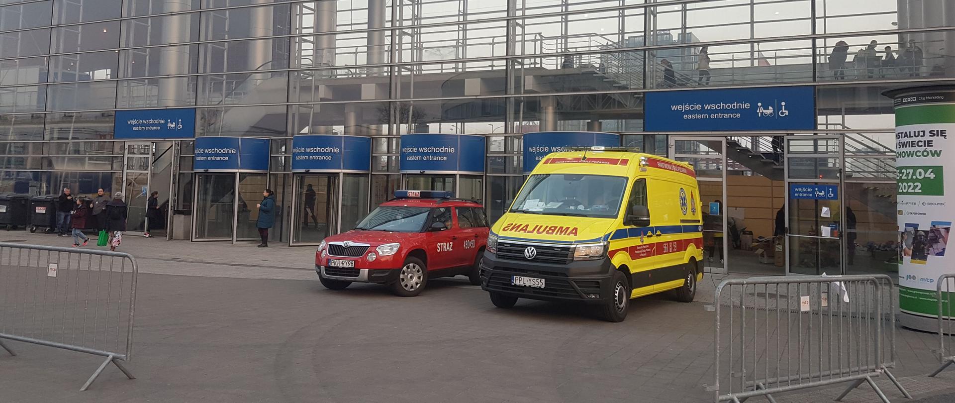 Przed halą na międzynarodowych targach poznańskich stoi karetka w kolorze żółtym oraz samochód operacyjny krotoszyńskiej komendy. W oddali widać ludzi przemieszczających się po placu