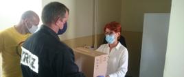 Pani dyrektor szkoły zabezpieczona w maskę odbiera pudło ze środkami do dezynfekcji. Pudło wręczane przez strażaka w umundurowaniu koszarowym, zabezpieczonego w maskę oraz rękawiczki lateksowe. Po lewej stronie zdjęcia postronna osoba w żółtej koszulce.