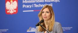 Wiceminister rozwoju, pracy i technologii Anna Kornecka stojąca przy mównicy na konferencji prasowej, z tyłu na jej plecami baner z logo MRPiT i napisem "Plan dla Pracy i Rozwoju"
