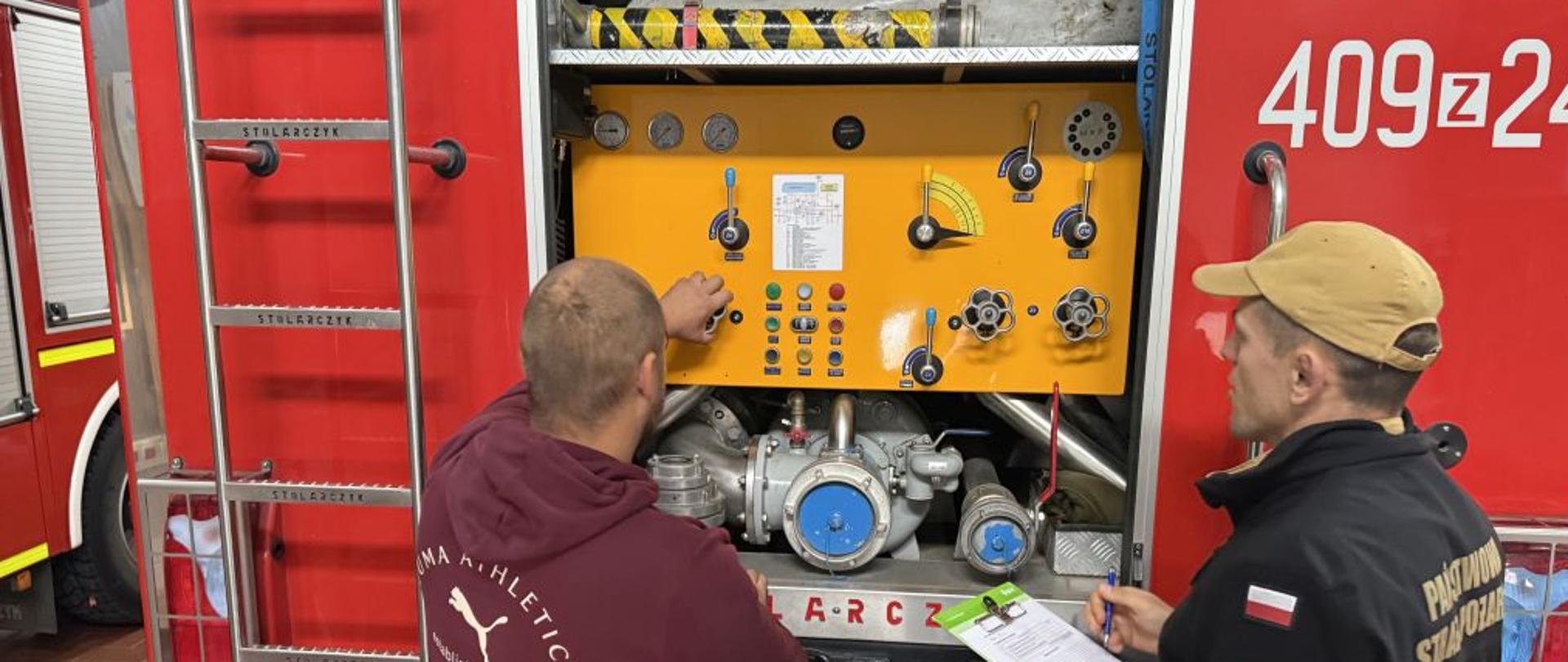 Dwóch strażaków prowadzi kontrolę autopompy wozu strażackiego. Jeden z nich testuje działanie panelu sterowania, drugi sporządza notatki kontrolne. W tle widoczne są oznaczenia pojazdu pożarniczego. Sprawdzenie obejmuje działanie układu wodnego i ciśnieniowego. Kontrola przebiega w spokojnej, roboczej atmosferze.