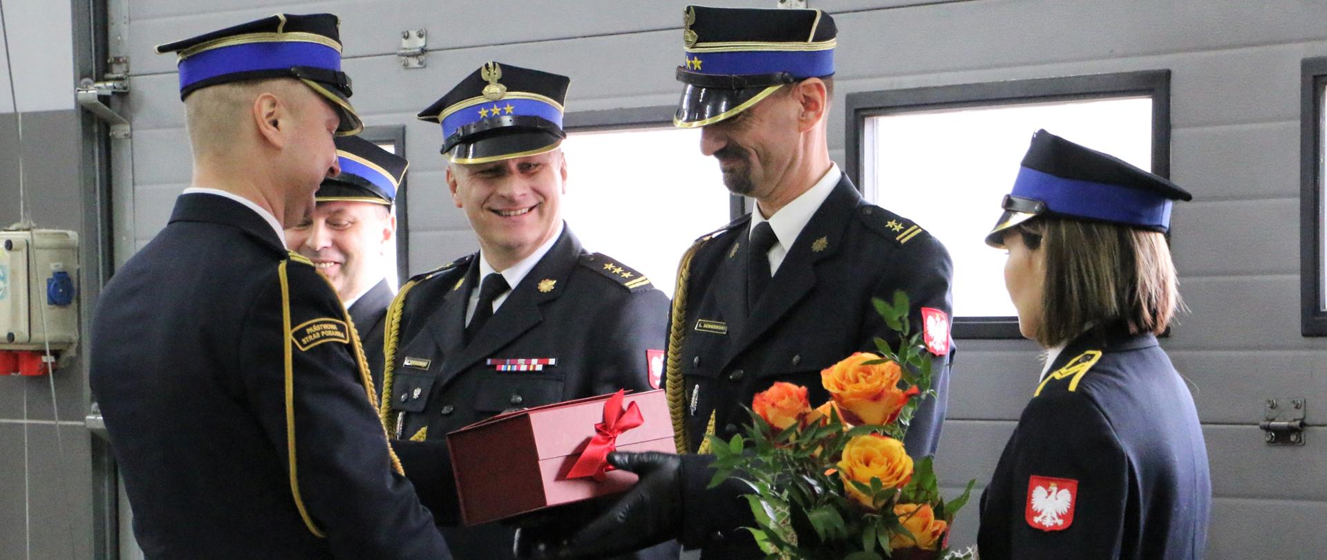 Komendant powiatowy wręcza prezent odchodzącemu ze służby zastępcy. Obok stoi przełożony, za nim następca. Z prawej strony funkcjonariuszka podaje kwiaty.