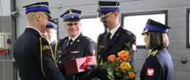 Komendant powiatowy wręcza prezent odchodzącemu ze służby zastępcy. Obok stoi przełożony, za nim następca. Z prawej strony funkcjonariuszka podaje kwiaty.
