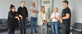 Szkolenie z pierwszej pomocy. Świetlica Komendy Powiatowej Policji w Rawiczu. Pięciu uczestników szkolenia (3 kobiety i 2 mężczyzn) słucha i obserwuje czynności wykonywane przez strażaka. Ten trzyma w rękach manekina "niemowlaka" . W tle drzwi do pomieszczenia, a na ścianie zawieszony jest klimatyzator.