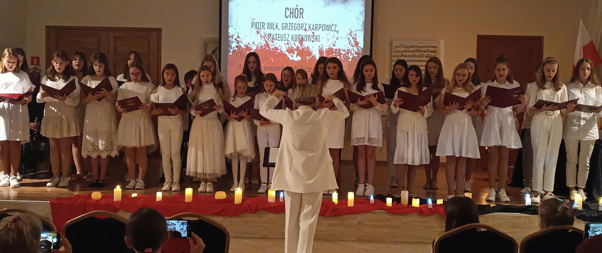 Zdjęcie przedstawia chór śpiewający pieśni patriotyczne. Chór jest ubrany na biało.W tle na ekranie widnieje napis chór, oraz skład zespołu nauczycieli akompaniujących: Piotr Wilk, Grzegorz Karpowicz, Mateusz Kurkowski. Ekran jest w kolorze biało-czerwonym. Dalej widzimy grafikę przedstawiającą partyturę, brązowe drzwi, oraz stół, zza którego wystaje biało-czerwona flaga. Przed chórem na podłodze leży czerwony materiał, a na nim białe zapalone świece. Przed sceną stoi Pani Ewelina Chrzanowska i dyryguje chórem. Po bokach widać publiczność słuchającą koncertu. 