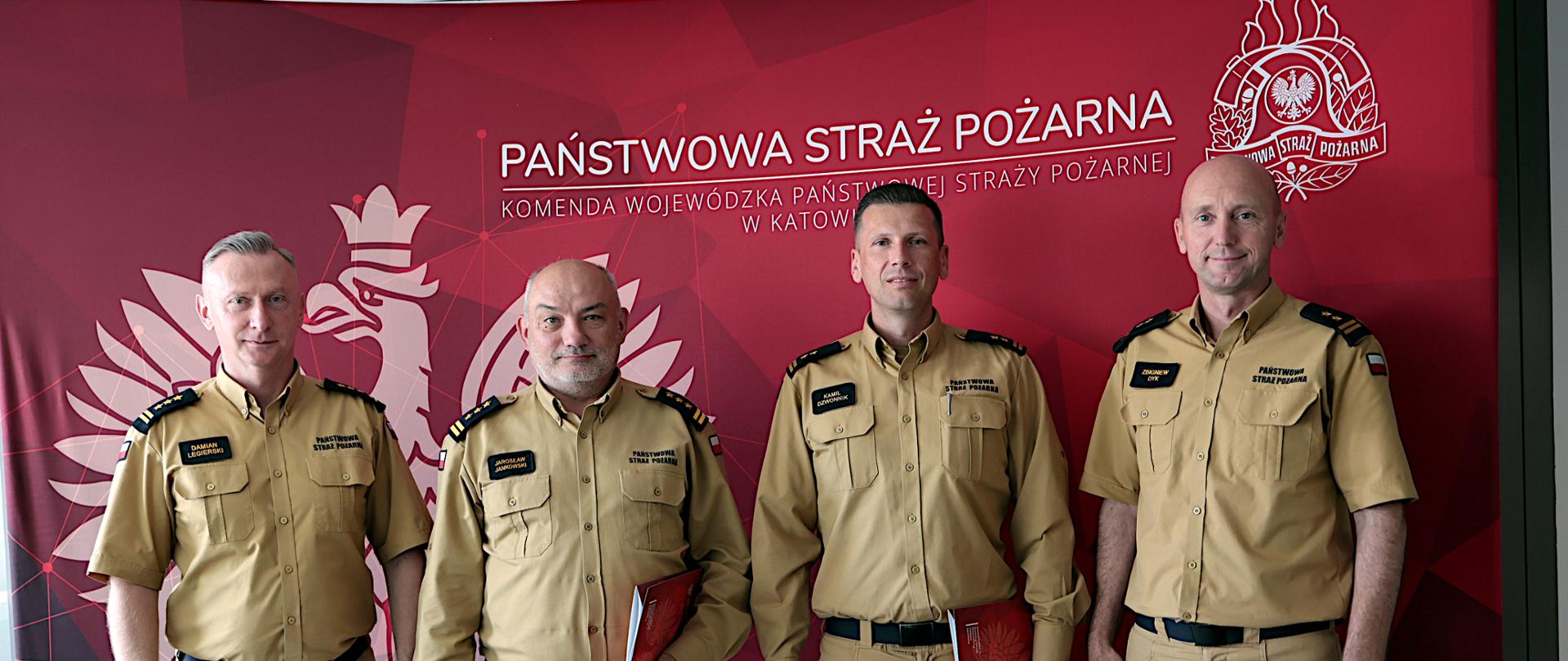 Na zdjęciu czterech funkcjonariuszy Państwowej Straży Pożarnej stoi obok siebie na tle czerwonej ścianki z dużym godłem i napisem Komendy Wojewódzkiej Państwowej Straży Pożarnej w Katowicach. Od lewej strony śląski komendant wojewódzki Państwowej Straży Pożarnej, st. bryg. Damiana Legierskiego, komendant Centralnej Szkoły Państwowej Straży Pożarnej w Częstochowie, st. bryg. Jarosław Jankowski, komendant miejski Państwowej Straży Pożarnej w Częstochowie, bryg. Kamil Dzwonnik oraz zastępca śląskiego komendanta bryg. Zbigniew Dyk. Wszyscy ubrani są w beżowe mundury służbowe i patrzą w stronę aparatu. Dwóch strażaków stojących pośrodku trzyma czerwone teczki z dokumentami.