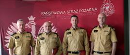 Na zdjęciu czterech funkcjonariuszy Państwowej Straży Pożarnej stoi obok siebie na tle czerwonej ścianki z dużym godłem i napisem Komendy Wojewódzkiej Państwowej Straży Pożarnej w Katowicach. Od lewej strony śląski komendant wojewódzki Państwowej Straży Pożarnej, st. bryg. Damiana Legierskiego, komendant Centralnej Szkoły Państwowej Straży Pożarnej w Częstochowie, st. bryg. Jarosław Jankowski, komendant miejski Państwowej Straży Pożarnej w Częstochowie, bryg. Kamil Dzwonnik oraz zastępca śląskiego komendanta bryg. Zbigniew Dyk. Wszyscy ubrani są w beżowe mundury służbowe i patrzą w stronę aparatu. Dwóch strażaków stojących pośrodku trzyma czerwone teczki z dokumentami.
