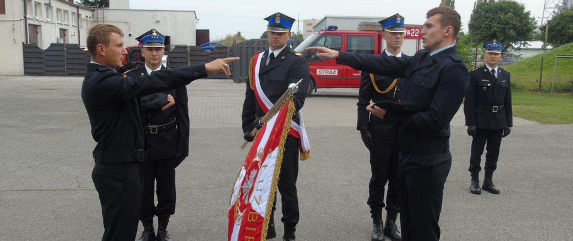 Na placu przed garażami, poczet sztandarowy składający się z trzech strażaków ubranych w umundurowanie wyjściowe, na głowach mają założone rogatywki. Jeden ze strażaków trzyma sztandar komendy skierowany do przodu. Po obu stronach sztandaru stoją strażacy ( dwóch strażaków) ubrani w mundury służbowe. Mają wyciągniętą prawą rękę do przodu w kierunku sztandary, w lewych trzymają nakrycia głowy ( berety). składają ślubowanie na sztandar jednostki.