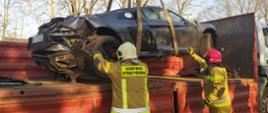 Kolizja drogowa-dachowanie audi. Droga gminna Rawicz-Przywsie. Dwóch strażaków pomaga przy załadunku rozbitego audi na samochód ciężarowe. Auto ładowane jest za pomocą HDS i pasów transportowych. W tle drzewa.