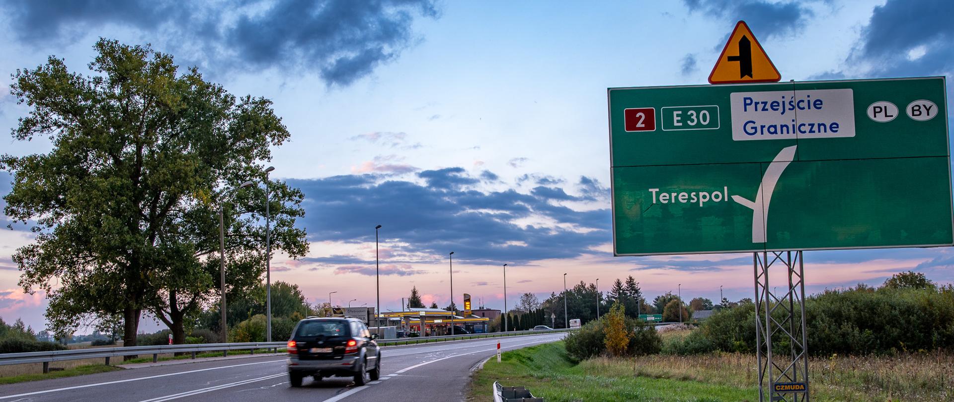Zdjęcie przedstawia jezdnię i samochód. W tle widać stację benzynową. Po prawej stronie widać dużą, zieloną tablicę wskazującą drogę na Terespol oraz na Przejście Graniczne.