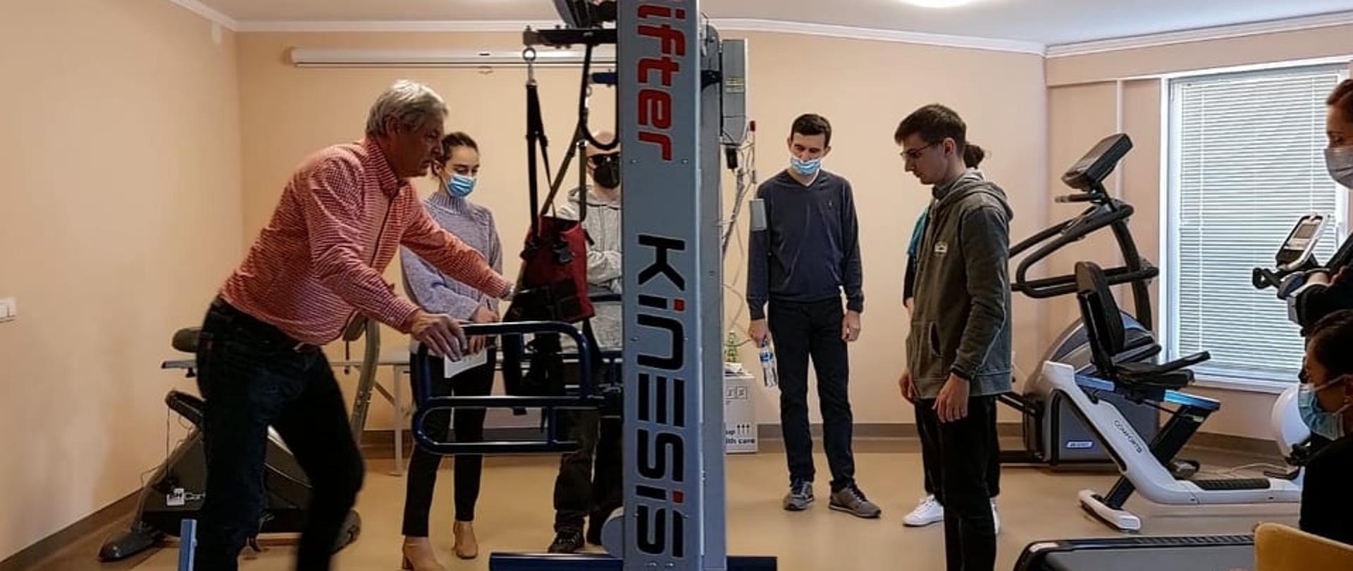 Osiem osób stoi w sali wokół urządzenia do rehabilitacji. Jedna z nich, mężczyzna, prezentuje jak używa się urządzenia.