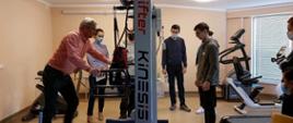 Osiem osób stoi wokół sprzętu do rehabilitacji. Jedna z nich, mężczyzna, prezentuje jak korzystać z urządzenia.