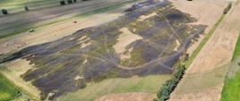 Zdjęcie lotnicze przedstawia rozległy obszar pól uprawnych z dużym pogorzeliskiem pośrodku, na którym widoczne są ślady kół pojazdów. Wypalona ziemia tworzy ciemną, nieregularną plamę otoczoną nienaruszonymi, jaśniejszymi łanami zbóż oraz zielonymi łąkami. W oddali, na sąsiednich polach, pracują maszyny rolnicze, w tym czerwony kombajn, a na horyzoncie rozciąga się panorama wiejskiej okolicy z kępami drzew i zabudowaniami.