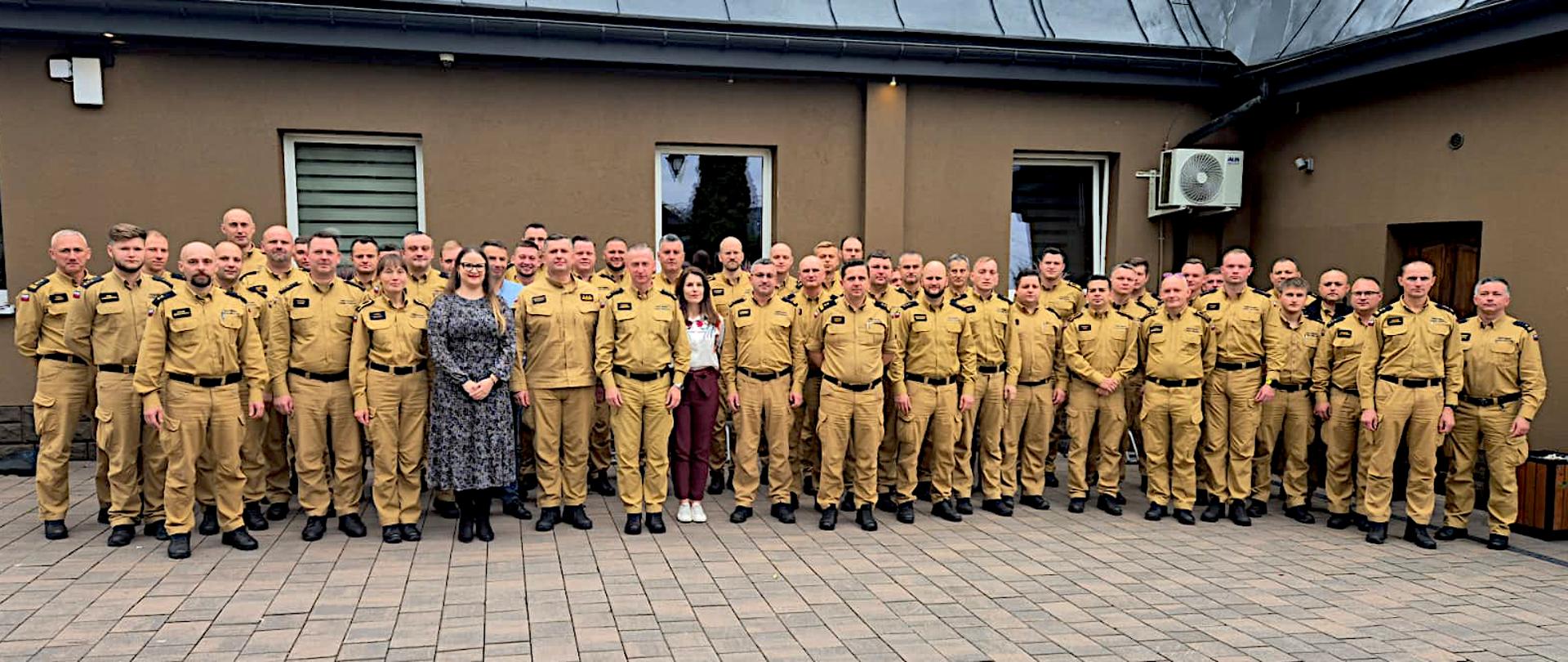 Na zdjęciu stoi duża grupa osób – około czterdziestu kilku mężczyzn i dwóch kobiet – ustawionych w kilku rzędach przed budynkiem o brązowych ścianach. Większość osób to strażacy ubrani w jednakowe, piaskowe mundury służbowe z czarnymi pagonami i naszywkami na piersi. Stoją w równym szeregu, patrząc w stronę aparatu. Wśród nich, w środkowej części zdjęcia, stoją dwie kobiety w cywilnych ubraniach. Tło stanowi ściana budynku z dwoma oknami i drzwiami. Zdjęcie grupowe wykonane na zewnątrz.