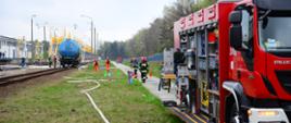 Samochód pożarniczy z otwartymi skrytkami. N 2 planie widoczny wagon cysterna do transportu substancji niebezpiecznych, wagon stoi na bocznicy kolejowej. W oddali widoczni są strażacy prowadzący działania ratownicze. W okolice wagonu rozwinięte zostały węże pożarnicze.