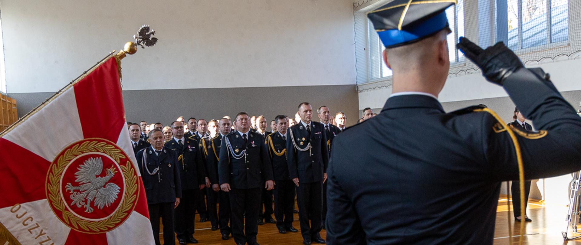 Funkcjonariusz PSP salutuje przed sztandarem z napisem „Ojczyzny” podczas uroczystości Święta Niepodległości w Opolu. W tle widoczni mundurowi w szyku oraz dokumentujący wydarzenie fotografowie.